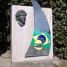 Photo d'une monument à la mémoire de Senna sur le circuit de Catalogne à Montmeló formé d'un rectangle de pierre avec le vosage de Senna, son casque et le drapeau du Brésil.