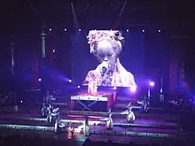 Photo d'une scène de concert avec des chorégraphes disposés en « V », l'artiste sur une plateforme au milieu et un écran géant à l'arrière plan, projetant l'image de la chanteuse