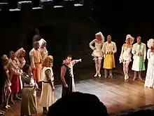 Photographie d'une scène de théâtre où deux groupes d'acteurs costumés de façon médiévales se font face. Au milieu d'eux un homme montre le groupe de droite de façon accusatrice.