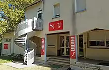 Photographie en couleur de l'entrée d'un bâtiment avec un escalier externe vu de trois-quarts face.