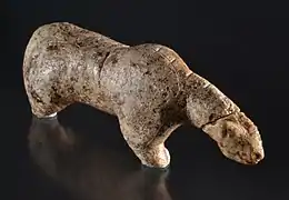 Photographie en couleurs sur fond noir et vue du dessus d'une statuette en ivoire représentant un ours des cavernes, ses pattes amputées reposant sur un socle de verre.