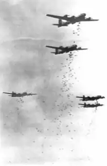 Photographie en noir et blanc d'une formation de cinq quadrimoteurs. Chacun d'eux largue plusieurs dizaines de bombes.