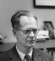 Photographie en noir et blanc du psychologue américain Burrhus Frederic Skinner, auteur de « Walden Two » (1948). L'avant du crâne légèrement dégarni et portant des lunettes, B. F. Skinner est habillé d'un costume et d'une cravate. Derrière lui, une étagère de livres.
