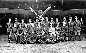 Photographie noir et blanc. Vingt-cinq hommes en uniforme, debout ou assis, devant un biplan avec une hélice à quatre pales.