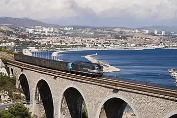  Un train passe sur un viaduc, à l'arrière plan un littoral urbanisé et la mer.