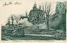 Carte postale en noir et blanc : une locomotive à vapeur avec son panache de fumée, devant le bâtiment de la gare ; figurent deux mots manuscrits : « Amitié Henri ».
