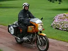 Gold BMW R90S motorcycle ridden through a park by a rider in black waterproof one piece suite