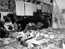 photo en noir et blanc d'une foule marchant dans une allée, au premier plan des revues sous plastique
