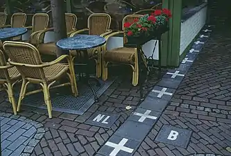Ligne de pavés gris surmontés de croix blanches, qui délimite la frontière au pied d'un café.