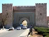 Route passant sous une porte monumentale à arc outre passé, flanquée de deux imposantes tours symétriques, et se poursuivant sur une muraille.