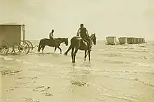 2 chevaux montés, dont 1 tire  une cabine cubique à 4 roues vers une rangée de cabines semblables déjà placées au large de la plage.