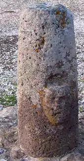 Un bétyle de pierre claire en forme de petit pilier, avec un visage sculpté en relief.