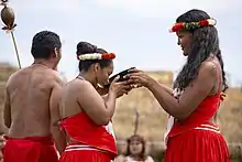 Deux femmes, un homme, vêtus de sortes de pagnes rouges. Une femme boit dans un grand bol.