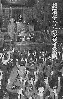 Photo en noir et blanc de la salle des représentants avec, au premier plan, les membres de la chambre debout devant leurs sièges, les bras levés dans le geste du Banzai