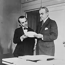 Deux hommes en costume, de face, debouts derrière un bureau, regardant un objet ovale et plat qu'ils tiennent chacun d'une main.