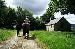 Cavalière sur une jument de trait, vue de dos, près d'une chapelle.