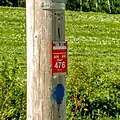 Une pièce de plastique rouge carrée portant l'inscription « GR A1 SOS 911 point 476 SIA/IAT » en blanc, fixée sur un poteau téléphonique