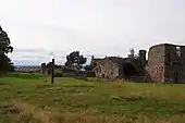 Photographie d'un champ dans lequel se dresse une grande croix de bois. Derrière celle-ci sont placées les ruines médiévales d'un monastère.
