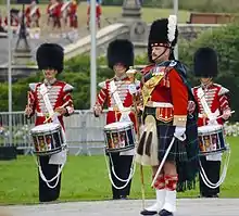 Photographie de trois musiciens militaires en uniforme composé d'une tunique rouge, d'un pantalon noir et d'un bonnet de poil noir jouant sur des tambours ainsi que de leur dirigeant portant un uniforme composé d'une tunique rouge, d'un kilt et d'un bonnet de poil noir comprenant une plume blanche tenant un bâton dans sa main droite et une épée dans sa main gauche