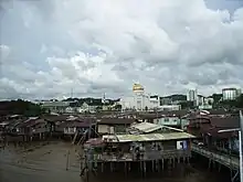 Photographie d'un quartier pauvre de la capitale, Bandar Seri Begawan. Au premier plan se trouvent des maisons sur pilotis délabrées. En arrière-plan se trouve la mosquée Omar Ali Saifuddin et des immeubles modernes en béton.