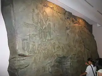 Photographie d'une bloc de pierre d'environ 4 mètres de haut par 8 mètres de large, dans un musée, sur lequel des gravures sont visibles sur sa face plane.
