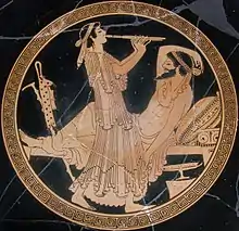 Tondo doré sur céramique noire représentant un homme barbu allongée sur une banquette qui écoute un joueur de flute debout.