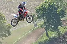 Une moto "saute" au dessus d'un chemin, des arbres, des vignes.