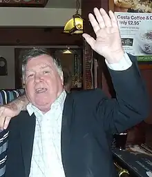 Portrait de face d'un homme debout dans un pub la main gauche levée, un fan le coude appuyé sur son épaule droite.