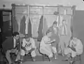 Deux joueurs des Royaux de Montréal et un joueur des Dodgers de Brooklyn assis sur des tabourets dans le vestiaire, avec trois autres hommes en costume, en 1946.