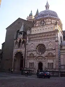 Image photographique d'une façade de forme carrée avec au centre un portail surmonté d'une grande rosace. La façade est enrichie de motifs picturaux, avec l'utilisation d'une trichromie blanc, rose, violet et le motif des losanges, surmontée d'une coupole.