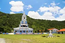 Photo en couleur montrant un bâtiment blanc et bleu avec un clocher au milieu. Devant se trouve une grande pelouse sur laquelle se trouve une croix blanche ; en arrière-plan, la forêt et quelques maisons.