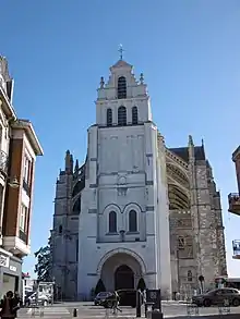 Tour-porche présentant une couleur très blanche. En haut de la tour, une croix surmontée d'un coq dorée et, un peu plus bas, deux têtes sculptées de saint Quentin avec les clous sur chaque épaule. Ciel bleu.
