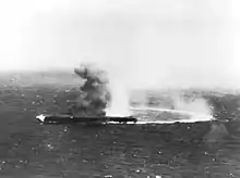 Une haute colonne de fumée s'élève au-dessus d'un porte-avions tandis qu'une large lueur est visible à sa proue. Une trainée derrière le navire indique qu'il a violemment viré sur tribord