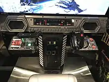 Sorte de meuble gris et noir imitant un cockpit de vaisseau, avec des boutons et des lumières, une manette de jeu de chaque côté et un monnayeur au centre.