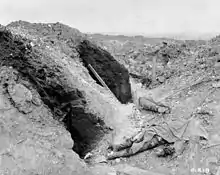 Photographie en noir et blanc de cadavres étendues dans une tranchée détruite avec un soldat se tenant debout en arrière-plan