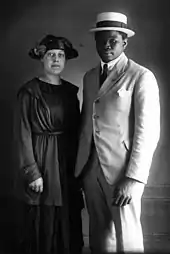 Un couple avec un homme en costume blanc et une femme habillée d'un robe et un gilet. Tous deux portent un chapeau.
