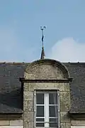 Photographie d'une lucarne surmontée d’une girouette, sur fond de toit en ardoise.