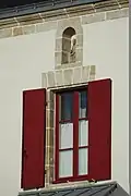 Photographie d'une fenêtre aux volets rouges, surmontée d’une niche contenant une statuette.