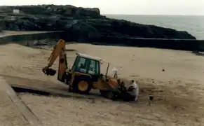 Photographie d’une pelleteuse orange s’avançant sur une plage, avec deux hommes en blanc.