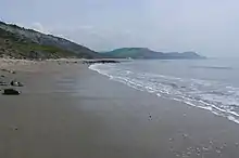 Vaste plage de sable gris, léchée à droite par les vagues. Arrière plan de collines s'abaissant vers la mer