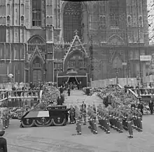le cercueil de la reine est posé sur un affût de canon drapé du drapeau belge et tiré par des militaires devant la façade de la cathédrale recouverte en partie d'échafaudages et dont l'entrée en ornée d'un dais de tissu sombre
