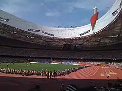 Vue de l'intérieur d'un stade avec les pistes d'athlétisme en rouge entourant un stade de football en vert, et de hautes tribunes remplies de spectateurs