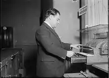 Photographie en noir et blanc d'une personne debout, en costume de couleur sombre, trois-quart de profil, regardant et actionnant une caisse enregistreuse