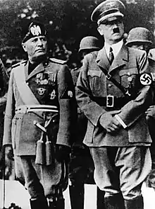 Photo noir et blanc montrant Benito Mussolini et Adolf Hitler, côte à côte, debout en uniforme, le regard fixé vers la gauche.