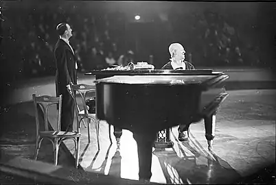 Grock et son partenaire, probablement Géo Lolé, en 1935 au cirque Busch (Berlin). Dans son numéro, Grock joue également un long moment au piano. Sur le piano est déposé un instrument à vent de la famille des cuivres, possiblement un cor ou un bugle, dont on distingue la bouche.