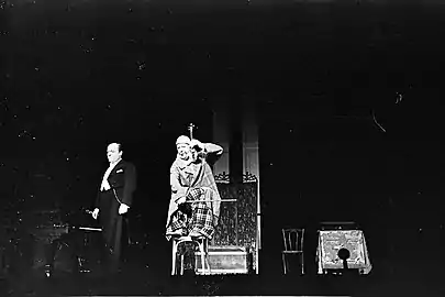 Grock et son partenaire Alfred Schatz à la Scala de Berlin en 1939. La photographie présente un passage classique du numéro où Grock monte sur une chaise avec un petit violon. En arrière plan, il est possible de distinguer le piano (sur la gauche) ainsi que d'autres accessoires : un paravent, un coffre, une autre chaise et une table.
