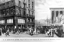Photo en noir et blanc d'un gros bâtiment d'angle à gauche, sur une place avec des passants, et des colonnes à droite