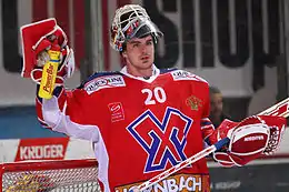 Photographie couleur d’un gardien de hockey sur glace, masque relevé sur sa tête, le bras droit levé, une gourde dans sa main.