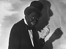 Photo en noir et blanc d'un homme en costume et au visage peint en noir tenant une cigarette.