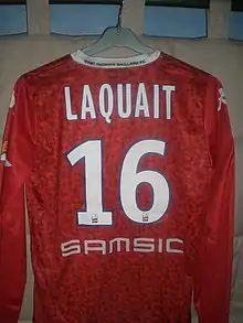 Photo montrant un maillot de football de couleur rouge aux longues manches avec un col blanc. Le dos du maillot est floqué "Laquait 16" en blanc avec contours bleu.